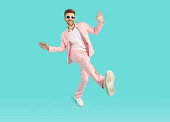 Wall Mural - Full body length shot of happy excited stylish young man dancing. Positive man in sunglasses and trendy light pink suit having fun while standing on one leg over isolated studio background
