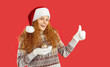 Girl is advertising Christmas sale or special offer. Portrait of cheerful cute young woman holding Christmas tree toy in her hand and showing thumb up. Woman in Santa hat isolated on red background.