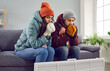 Young attractive couple are sitting on the sofa at home in warm clothes and warming their hands with heater in the living room, freezing from the cold. Heating problems, room is cold, people freezing.