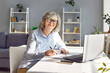 Senior smiling woman sitting on workplace at the desk working on laptop making notes planning budget. Concept of e-learning for mature people or distance freelance work career for retirement.