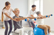 Coach or health care worker helping mature people to do sport exercises with dumbbells sitting on fit ball in gym. Physiotherapist helping retired man and woman in rehab in rehabilitation center.
