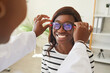 Optometrist puts new glasses on eyes of dark-skinned woman for testing and trying on. Joyful African American woman chooses glasses to improve vision. Optometry, eyewear and eye health concept.