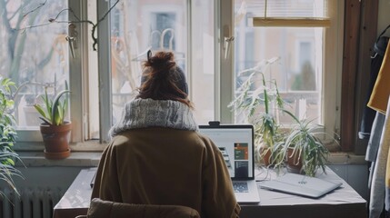 Poster - A young female uses her laptop computer to select and buy a new stylish coat at a clothing online web shop. The woman is sitting in a bright studio apartment with large windows in the daytime.