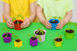Little kids hands planting tomato plants in colorful plastic pots on green table background. Closeup. Preparation for garden season. Children involvement in gardening. Front view.