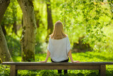 Fototapeta Tulipany - Young adult woman sitting on bench and staring at green leaves, grass and trees. Thinking about life at beautiful park in spring day. Spending time alone in nature. Peaceful atmosphere. Back view.