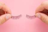 Fototapeta Tulipany - Young adult woman hand fingers holding and showing new dark black false lashes on light pink table background. Pastel color. Female beauty product. Closeup. Point of view shot. Top down view.