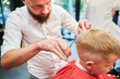 Man barber cutting little boy's hair using comb and scissors. Child getting haircut from adult male, likely barber. Professional hairdresser and cute client at modern barbershop.