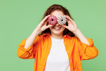 Wall Mural - Young smiling fun ginger woman wear orange shirt white t-shirt casual clothes hold in hand cover eyes with two donuts look camera isolated on plain pastel light green background. Lifestyle concept.