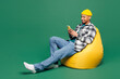 Full body young man of African American ethnicity he wear shirt blue t-shirt yellow hat sit in bag chair hold in hand use mobile cell phone isolated on plain green background studio Lifestyle concept