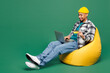 Full body young man wear shirt t-shirt yellow hat sit in bag chair using laptop pc computer hold credit bank card shopping online order delivery isolated on plain green background. Lifestyle concept.