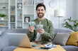 Cheerful young man holding US dollar bills with a laptop on his desk in a modern, well-decorated living room, exuding a sense of financial success and happiness.
