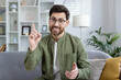 Cheerful young man in green shirt and glasses using hand gestures while speaking on a video call from a cozy, well-decorated living room.