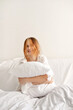 Good sleeping, time to sleep, youth daily health habit concept. Teenager girl sitting on bed, embracing pillow and smiling. White bedding, empty white wall background, candid emotions