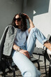 Fashionable beautiful curly woman with stylish sunglasses in fashion casual clothes with a blazer, white T-shirt and jeans sits and poses on the street in sunlight
