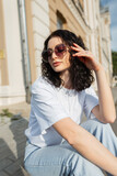 Fototapeta Panele - Stylish beautiful curly young girl model in fashionable clothes with a white T-shirt with heart-shaped sunglasses sits in the city
