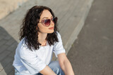 Fototapeta Panele - Stylish summer beautiful girl model with heart-shaped glasses in a white T-shirt sits on the asphalt in the city