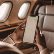 © Skip Monday - Close-up of Female Hands Holding Smartphone in Luxury Private Jet Cabin with Elegant Interior, Sunlit Atmosphere with Blank Screen phone mockup.