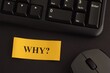 A paper note with the question Why? on it on a desk with a black keyboard and a grey wireless mouse. Close-up.