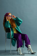 Fashionable redhead woman wearing purple headscarf, sunglasses, wide leg trousers, yellow blouse, green midi coat, silver ankle boots, posing on blue background. Studio full-length fashion portrait