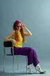 Fashionable redhead woman wearing purple headscarf, sunglasses, wide leg trousers, yellow blouse, silver ankle boots, posing on blue background. Studio full-length fashion portrait