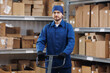 Service of factory logistic import export concept. Portrait man loader in blue uniform holding trolley with product on background warehouse.
