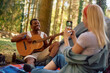 Happy black man playing acoustic guitar while camping with his girlfriend in woods.