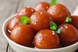 Gulab jamun, round balls with a sweet glaze and mint leaves on top in a white bowl. Close up view on a wooden table background. Indian traditional sweets, Asian food concept.