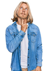 Wall Mural - Caucasian man with blond long hair wearing casual denim jacket thinking concentrated about doubt with finger on chin and looking up wondering