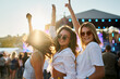 © artiemedvedev - Group of joyful young women dancing at sunlit beach music fest. Friends enjoy live concert, summer vibes, hands up in air. Trendy girls fun weekend on sandy shore with festival stage in background.