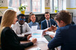 © artiemedvedev - Multiethnic colleagues discuss project in meeting at modern office. Caucasian woman holds graph chart, black businessman with colleague review data. Teamwork in corporate strategy session.
