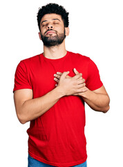 Wall Mural - Young arab man with beard wearing casual red t shirt smiling with hands on chest with closed eyes and grateful gesture on face. health concept.