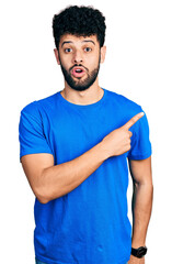 Poster - Young arab man with beard wearing casual blue t shirt surprised pointing with finger to the side, open mouth amazed expression.