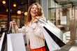 © maxbelchenko - Cheerful woman holding shopping bags. Beautiful woman in casual style shopping in the city. Consumerism, purchases, shopping, lifestyle, sale concept