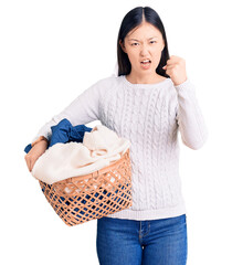 Sticker - Young beautiful chinese woman holding laundry basket with clothes annoyed and frustrated shouting with anger, yelling crazy with anger and hand raised