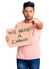 Wall Mural - Handsome latin american young man holding we need a change banner pointing with finger to the camera and to you, confident gesture looking serious