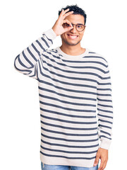 Canvas Print - Hispanic handsome young man wearing casual clothes and glasses doing ok gesture with hand smiling, eye looking through fingers with happy face.