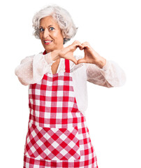 Wall Mural - Senior grey-haired woman wearing apron smiling in love showing heart symbol and shape with hands. romantic concept.