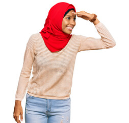 Wall Mural - Young african american woman wearing traditional islamic hijab scarf very happy and smiling looking far away with hand over head. searching concept.