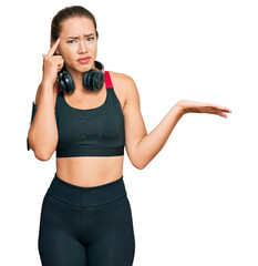 Beautiful blonde woman wearing gym clothes and using headphones confused and annoyed with open palm showing copy space and pointing finger to forehead. think about it.