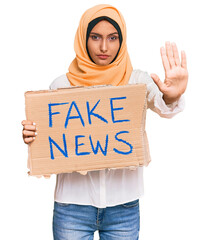 Poster - Young brunette arab woman holding fake news banner with open hand doing stop sign with serious and confident expression, defense gesture