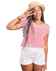 Wall Mural - Young african american woman with braids wearing summer hat doing peace symbol with fingers over face, smiling cheerful showing victory