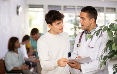 Wall Mural - Young male medic and visitor of clinic discussing something written on paper standing in waiting hall