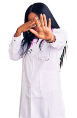 Wall Mural - Young african american woman wearing doctor stethoscope covering eyes with hands and doing stop gesture with sad and fear expression. embarrassed and negative concept.