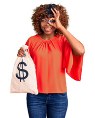 Canvas Print - Young african american woman holding money bag with dollar symbol smiling happy doing ok sign with hand on eye looking through fingers