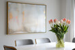A large framed painting with a gold leaf frame hangs above the dining table, the painting is beige and white abstract, the wall behind it is light grey, and tulips in vases on top of each side chair.