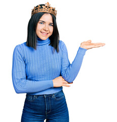 Poster - Young brunette woman with blue eyes wearing princess crown smiling cheerful presenting and pointing with palm of hand looking at the camera.