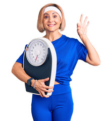 Canvas Print - Young blonde woman wearing sportswear holding weighing machine doing ok sign with fingers, smiling friendly gesturing excellent symbol