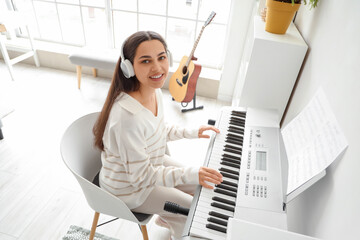 Wall Mural - Beautiful young happy woman in headphones listening music and playing synthesizer at home