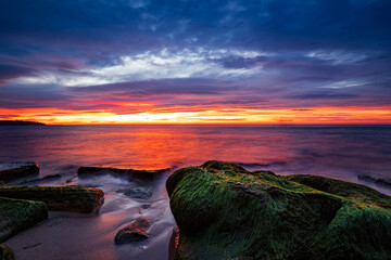 Wall Mural - Breathtaking sunset paints the sky above a tranquil sea, with moss-covered rocks in the foreground
