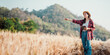 © Satori Studio - Curious young woman with notebook in hand pointing towards the horizon, standing amidst a vast wheat field with a mountain backdrop.